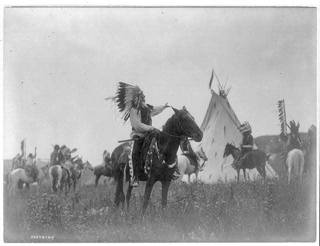 Dakota man, wearing war bonnet, sitting on horseback, his left hand outstreched toward tipi in background, others on horseback..jpg